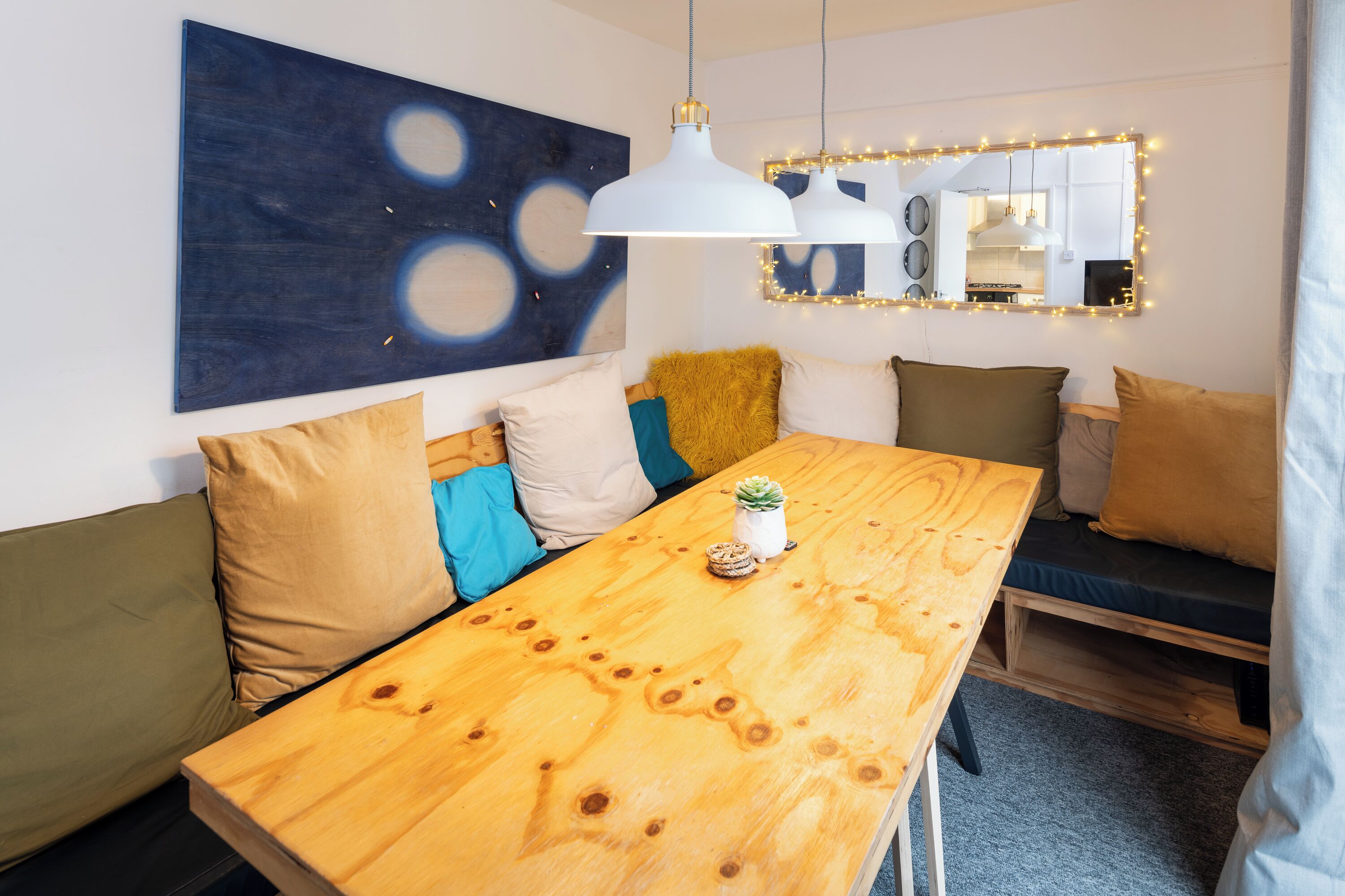 Un espace de vie moderne comprenant une table à manger en bois, un canapé avec des coussins, un tableau abstrait bleu et un miroir avec des lumières.