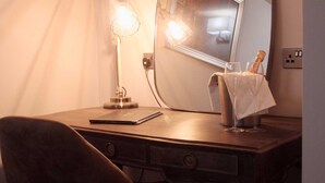 A wooden desk with a mirror, a lamp, a glass of water, and a bottle of champagne. - The Suffolk Hotel (Haverhill)