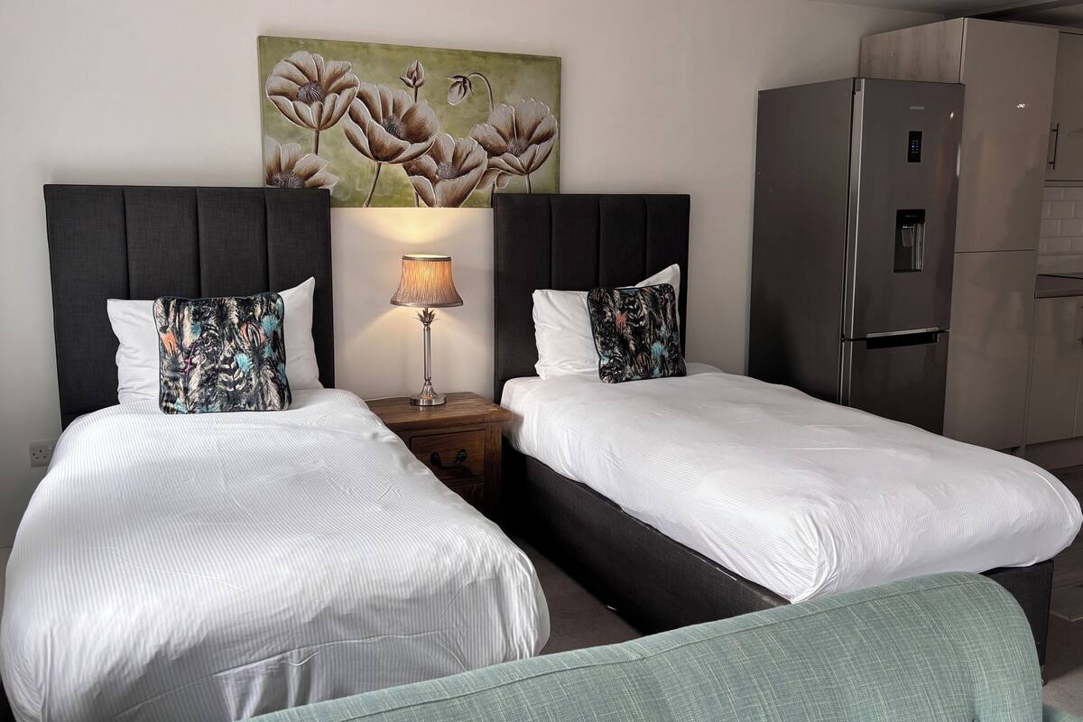 A hotel room with two beds, a nightstand with a lamp, a painting on the wall, and a kitchenette.