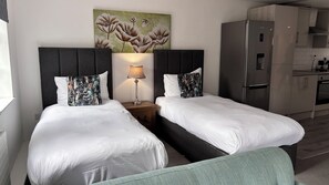 A hotel room with two beds, a nightstand with a lamp, a painting on the wall, and a kitchenette. - The Suffolk Hotel (Haverhill)