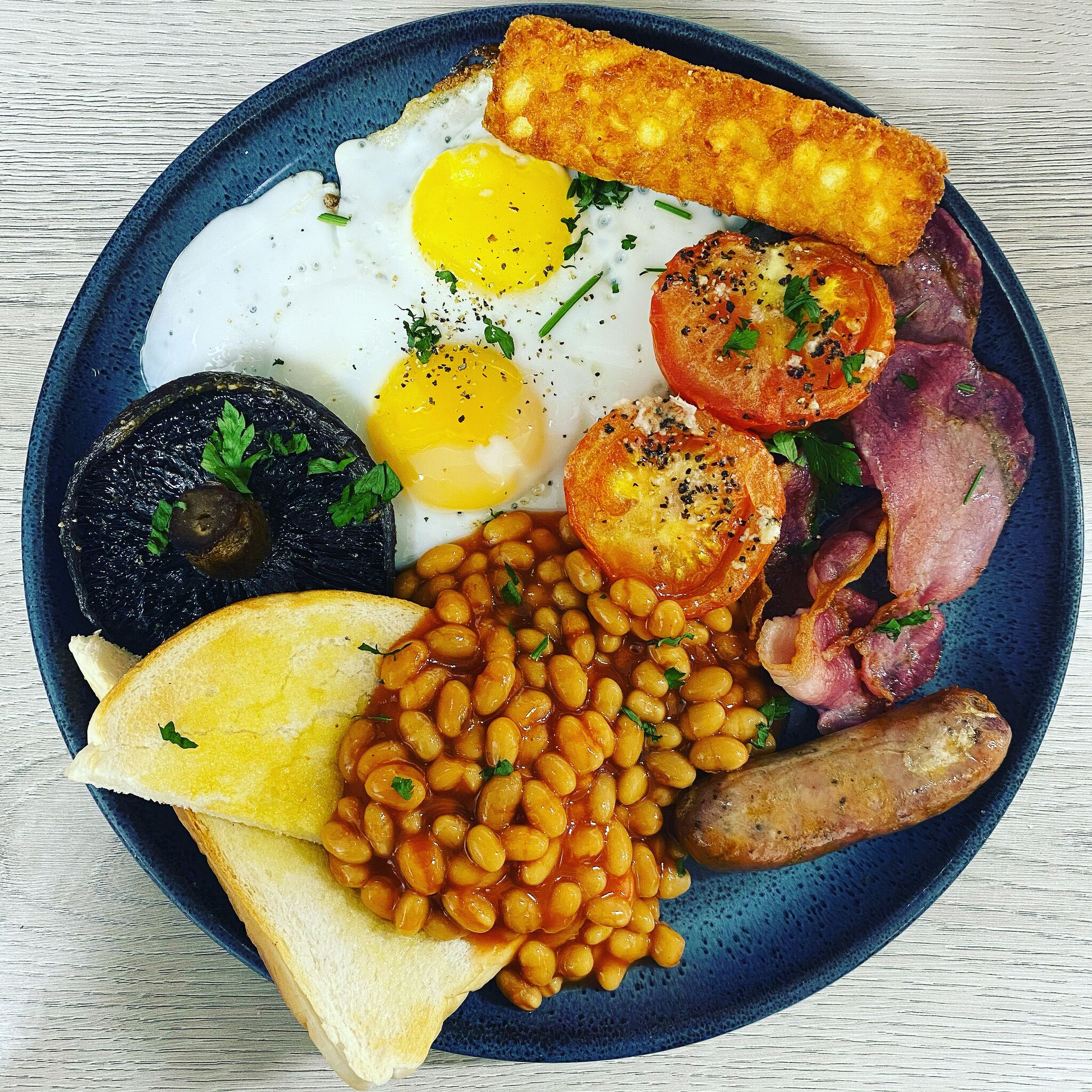 Un plato de comida para el desayuno que incluye huevos, tocino, salchichas, frijoles, tostadas y una papa al horno.