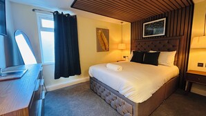 A hotel room with a bed, a desk, a chair, a window with curtains, a wall-mounted picture, and a lamp. - The Suffolk Hotel (Haverhill)