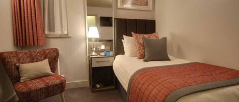 A hotel room with a bed, a chair, a nightstand, a lamp, and a painting on the wall.