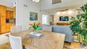 A modern kitchen with wooden cabinets, a dining area with a wooden table and wicker chairs, a living room with a sofa and a TV, and a large plant.