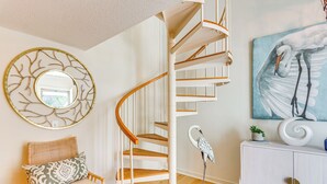 A spiral staircase with white railings and wooden steps, a round mirror with a tree design, a rattan chair with patterned cushions, a white cabinet with decorative items, and a painting of a bird on the wall.
