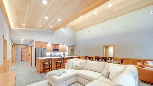 A spacious living area with a large L-shaped sofa, a wooden dining table, and a kitchen area with wooden cabinets and a refrigerator.