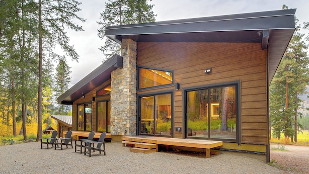 A modern cabin with a stone chimney, large windows, and a gravel patio with outdoor seating.