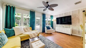 A living room with a yellow sofa, teal curtains, a ceiling fan, a white TV, and a wooden floor. - Tropical Pool Oasis on 1/4 Acre by Downtown Stuart (Stuart)