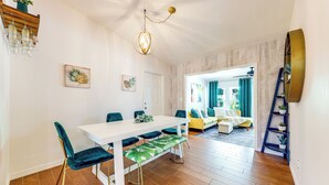 A dining area with a white table, green upholstered chairs, and a bench with a leaf pattern. There is a wooden shelf with a plant, a wall-mounted light fixture, and a door leading to another room. - Tropical Pool Oasis on 1/4 Acre by Downtown Stuart (Stuart)