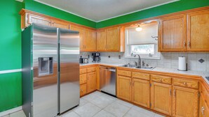 A kitchen with wooden cabinets, a stainless steel refrigerator, a dishwasher, and a window with blinds. - Cozy Home W/fire Pit: Drive to Palo Duro Canyon! (Amarillo)
