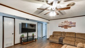 A living room with a flat-screen TV, a ceiling fan, a brown leather sofa, and a wooden floor. - Cozy Home W/fire Pit: Drive to Palo Duro Canyon! (Amarillo)