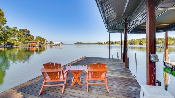 A wooden deck with two orange Adirondack chairs and a table, overlooking a calm lake with a clear sky. - Lake Keowee Home w/ Boat Dock in Seneca (Seneca)