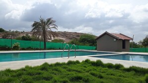 Une piscine dotée de deux échelles en métal, entourée de pelouse verdoyante et d’un petit bâtiment dont le toit est recouvert de tuiles rouges.
