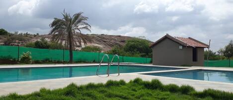 Una piscina con dos escaleras metálicas, rodeada de césped verde y un pequeño edificio con techo de tejas rojas.