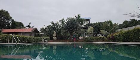 A swimming pool with a clear water surface, surrounded by lush greenery and a building with a red roof in the background.
