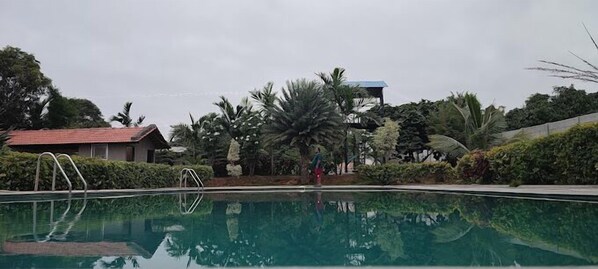 A swimming pool with a clear water surface, surrounded by lush greenery and a building with a red roof in the background.