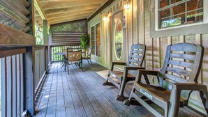Une véranda couverte avec un plancher en bois, deux chaises, une table et une fenêtre décorative.