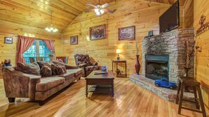 L’intérieur d’un chalet confortable, avec une cheminée en pierre, un plancher en bois, un canapé, une table basse et un téléviseur fixé au mur.