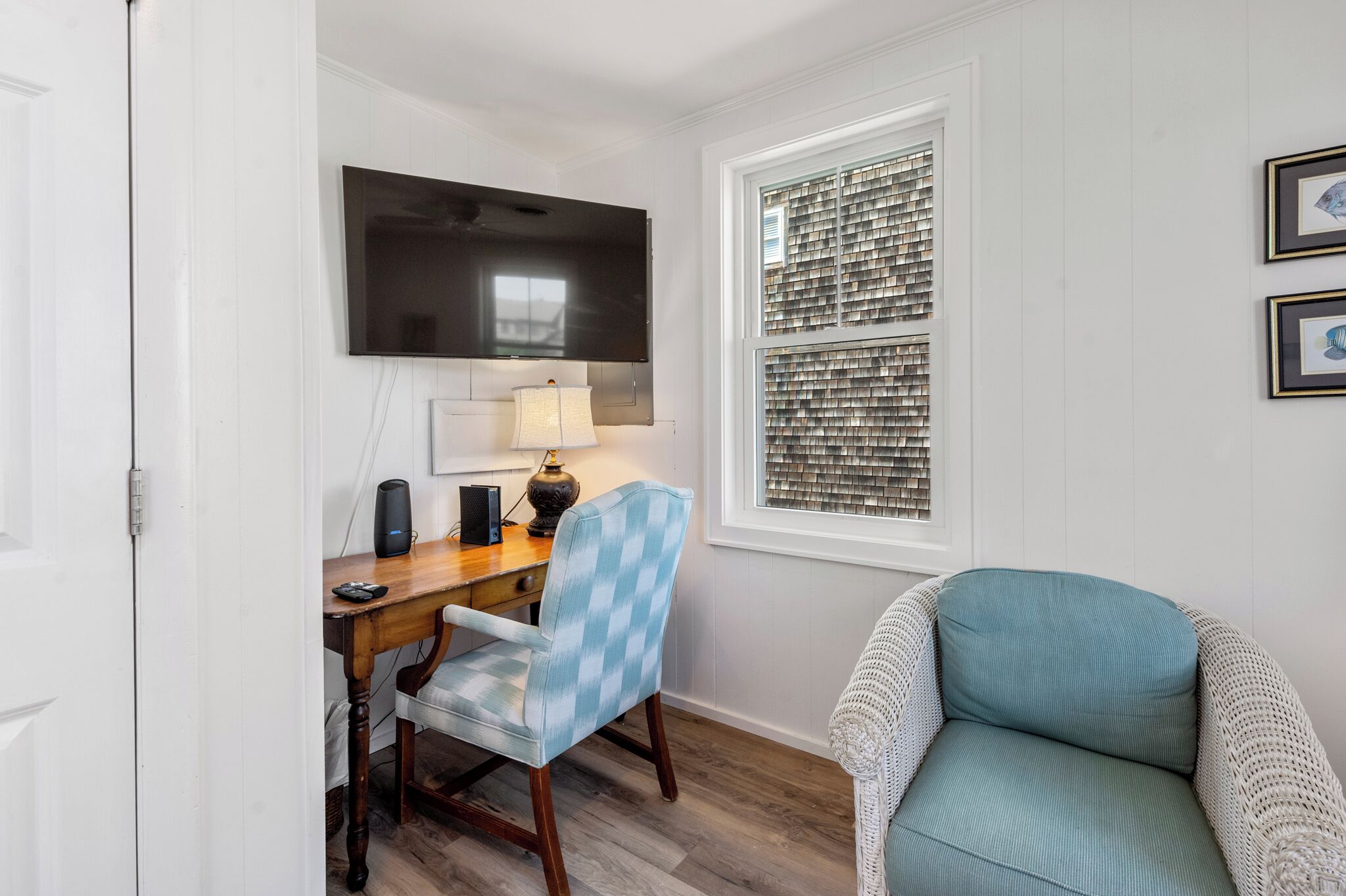 Une pièce avec un bureau en bois, une chaise à carreaux bleus, une chaise rembourrée de couleur turquoise, un téléviseur fixé au mur et une fenêtre avec des stores.