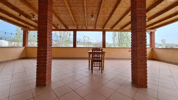 A spacious balcony with a tiled floor and wooden ceiling beams, featuring a single wooden chair and brick pillars. - Tirane Tranquility Cottage (Paskuqan)