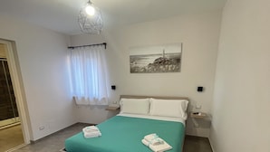 A neatly made bed with a teal bedspread and white pillows, a wall-mounted picture of a lighthouse, and a modern light fixture. - la casetta di zio e pia (Fiumicino)
