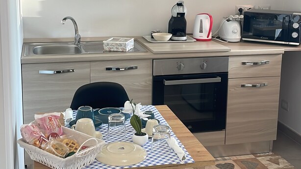 A modern kitchen with wooden cabinets, a stainless steel sink, a coffee maker, a toaster, and a microwave. There is a dining table with a checkered tablecloth and a basket of bread. - la casetta di zio e pia (Fiumicino)