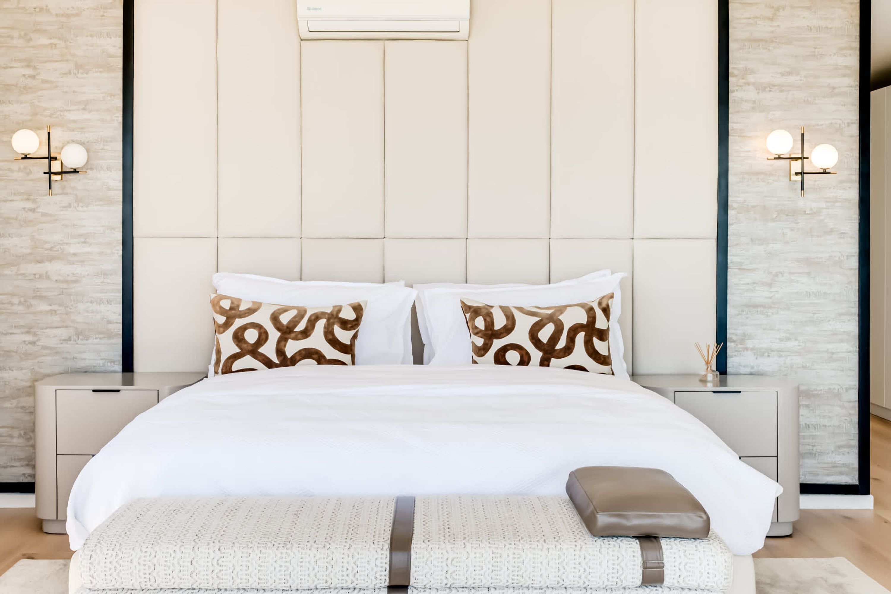 A modern bedroom with a large bed, two nightstands, a wall-mounted air conditioner, and two wall sconces.
