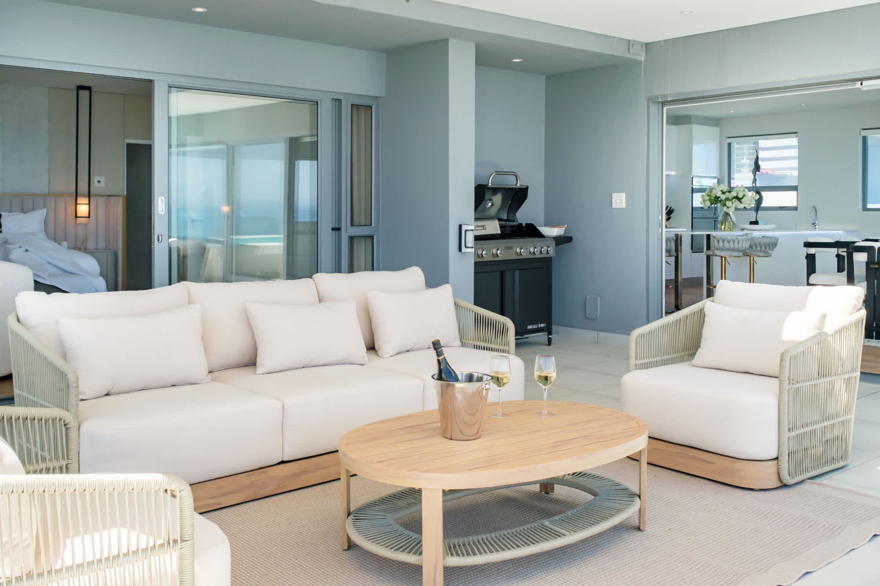 A modern living room with a white sofa, wicker chairs, a wooden coffee table, and a grill in the background.