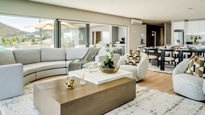 Ein modernes Wohnzimmer mit einem großen Ecksofa, einem Holztisch und einem Essbereich, wobei im Hintergrund eine Küche zu sehen ist.