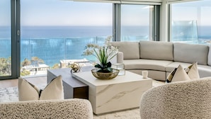 Ein modernes Wohnzimmer mit großem Fenster mit Meerblick, ein beiges Sofa, ein Marmortisch und eine Zierpflanze.