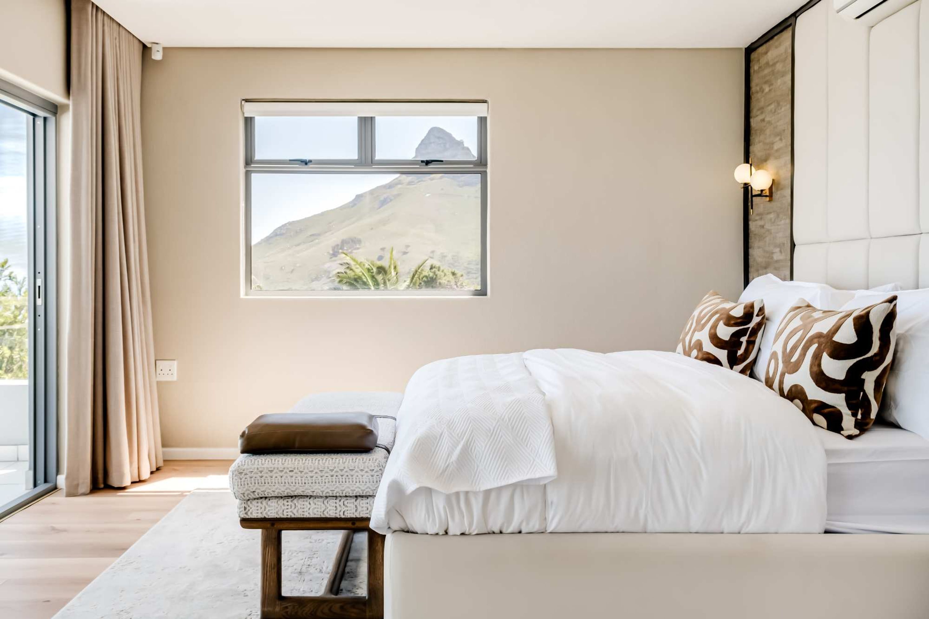 A bedroom with a large window, a bed with white bedding, a bedside table with a lamp, and a view of a mountain.