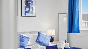 A hotel room with a bed, blue and white pillows, a blue blanket, a nightstand with a lamp, and a window with blue curtains. - Host Stay Stockenchurch (Milton Keynes)