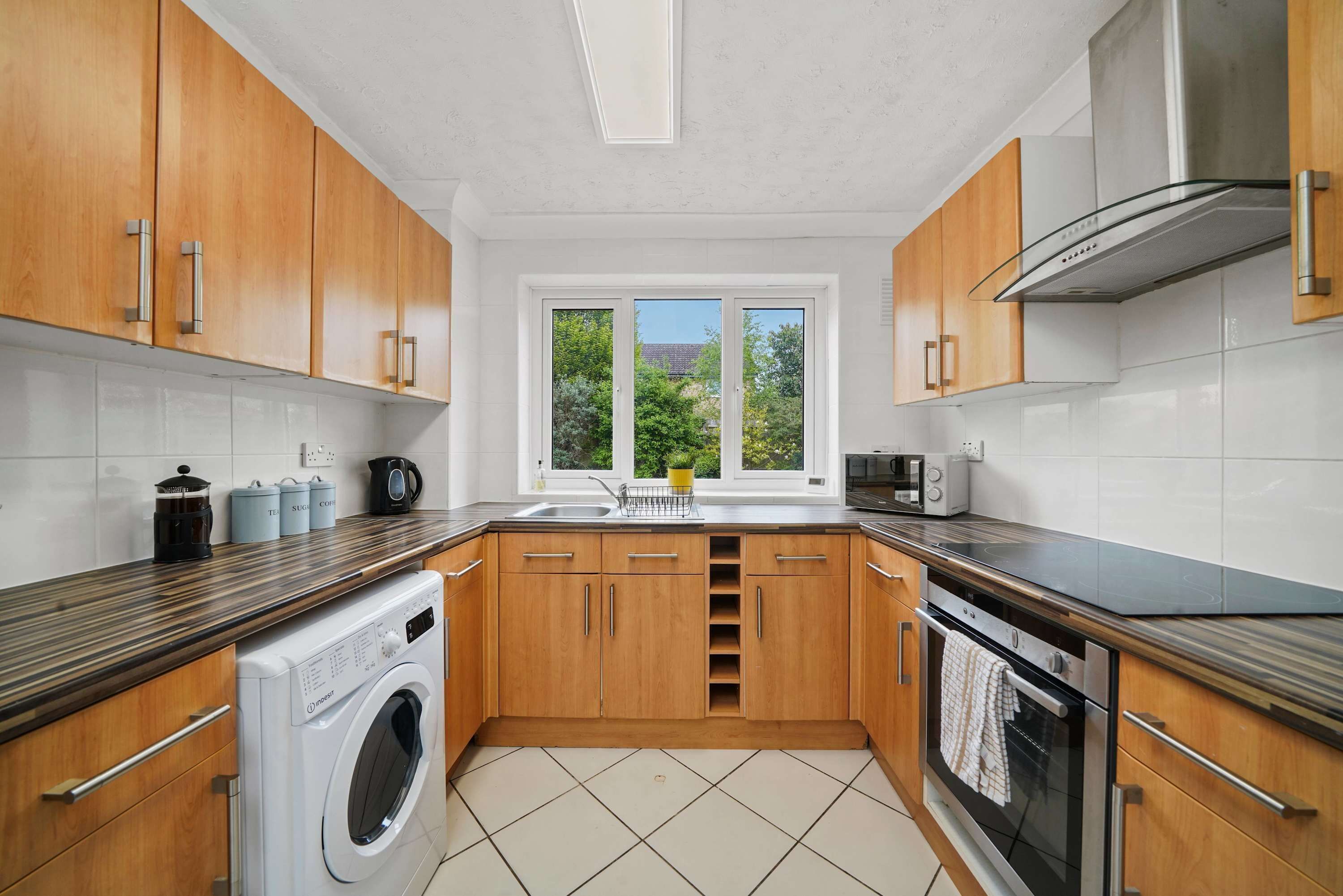 A modern kitchen with wooden cabinets, a built-in washing machine, a large window, and a stainless steel range hood.