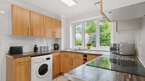 A modern kitchen with wooden cabinets, a built-in washing machine, a large window with a view of greenery, and a stainless steel countertop. - Host Stay Stockenchurch (Milton Keynes)