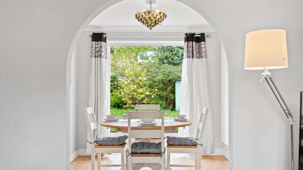 A dining area with a white table set for four, white chairs with grey cushions, a chandelier, and a lamp. - Host Stay Stockenchurch (Milton Keynes)