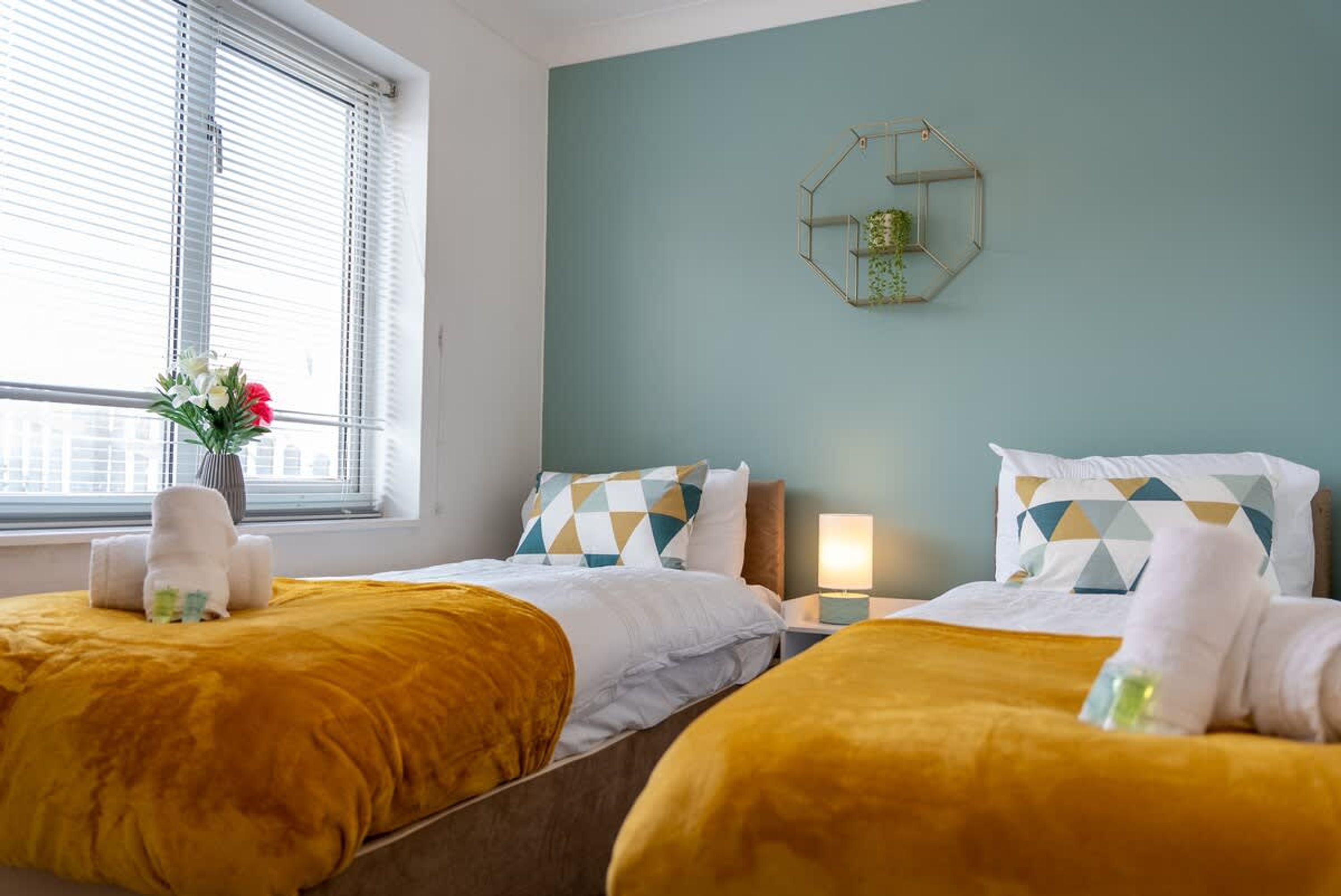 A hotel room with two beds, each with a yellow bedspread and white pillows with geometric patterns. There is a window with blinds, a bedside table with a lamp, and a wall-mounted hexagonal mirror with a plant.
