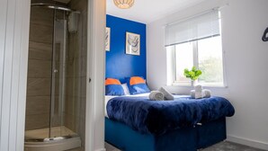 A bedroom with a glass-enclosed shower, a bed with blue bedding, and a window with blinds. - Host Stay Glazier Drive (Milton Keynes)