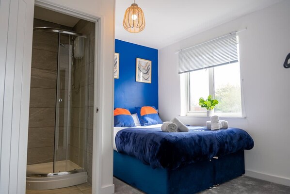 Une chambre avec une douche en verre, un lit avec une literie bleue et une fenêtre avec des stores.