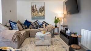 A modern living room with a sectional sofa, a coffee table, a painting, a TV, and a side table with a lamp. - Host Stay Glazier Drive (Milton Keynes)