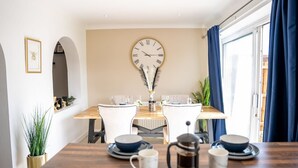 A dining area with a wooden table, white chairs, a clock on the wall, and a view of the outside through a window with blue curtains. - Host Stay Glazier Drive (Milton Keynes)