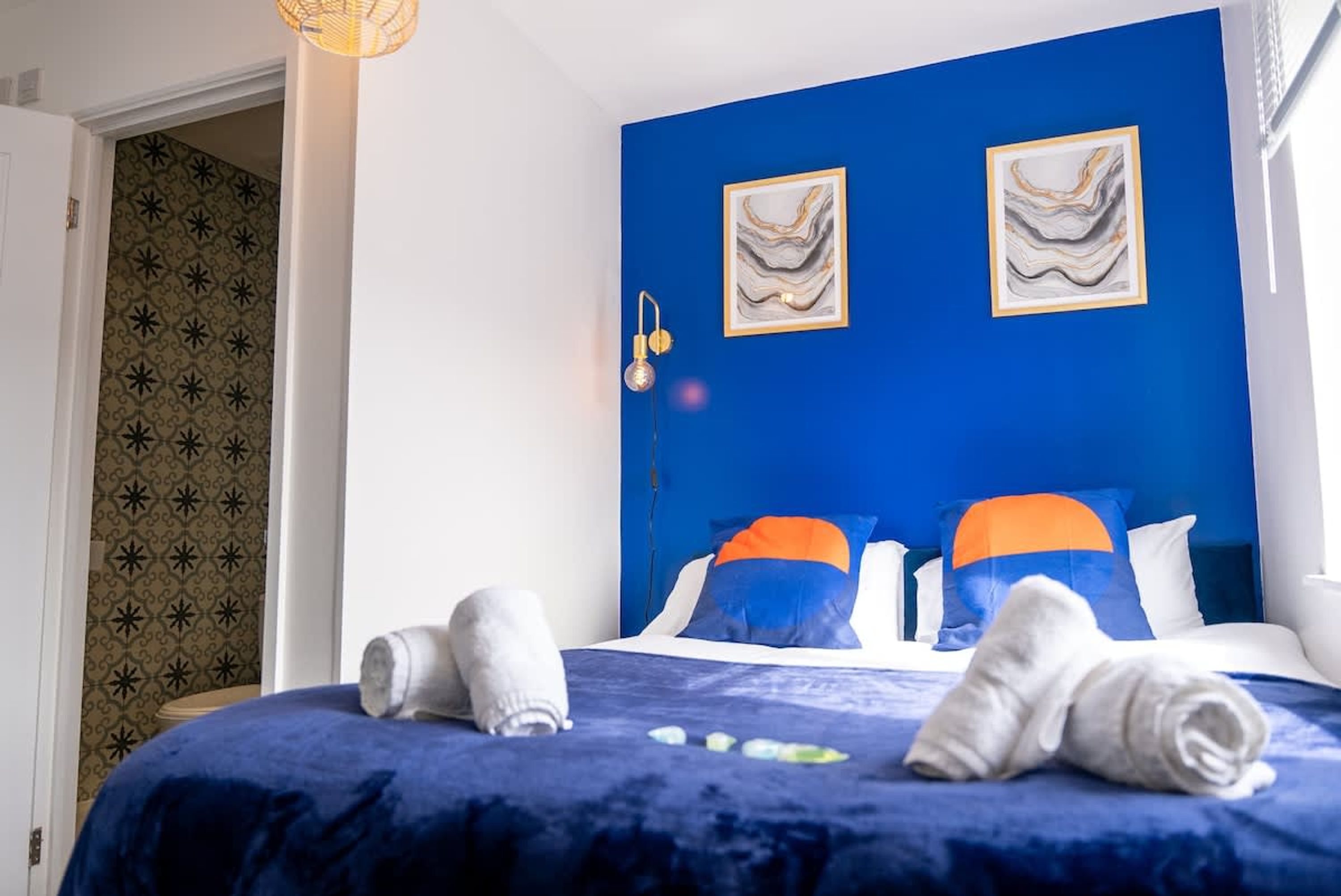 Habitación de hotel con una pared azul de acento, dos cuadros enmarcados, una cama con almohadas azules y anaranjadas, y una toalla sobre la cama.