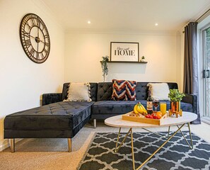 Una sala moderna con un sofá seccional, una mesa de centro redonda con frutas y bebidas, un reloj de pared y un cartel que dice 'Home sweet HOME'.