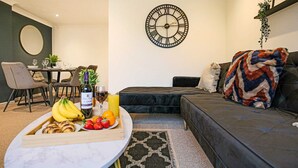 A modern living room with a dining area, a round table with fruits and drinks, a black sofa, and a large wall clock. - Host Stay Chicksands Avenue (Milton Keynes)