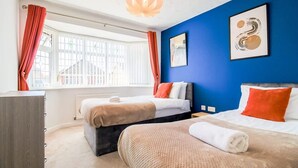 A hotel room with two beds, a dresser, a window with curtains, and a blue accent wall with artwork. - Host Stay Wiltshire Way (Milton Keynes)