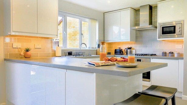 A modern kitchen with white cabinets, a large island with stools, a microwave, and a gas stove. - Host Stay Wiltshire Way (Milton Keynes)