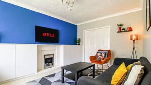 A living room with a blue wall, a fireplace, a black sofa with yellow pillows, a black coffee table, and a mounted TV displaying "NETFLIX". - Host Stay Wiltshire Way (Milton Keynes)