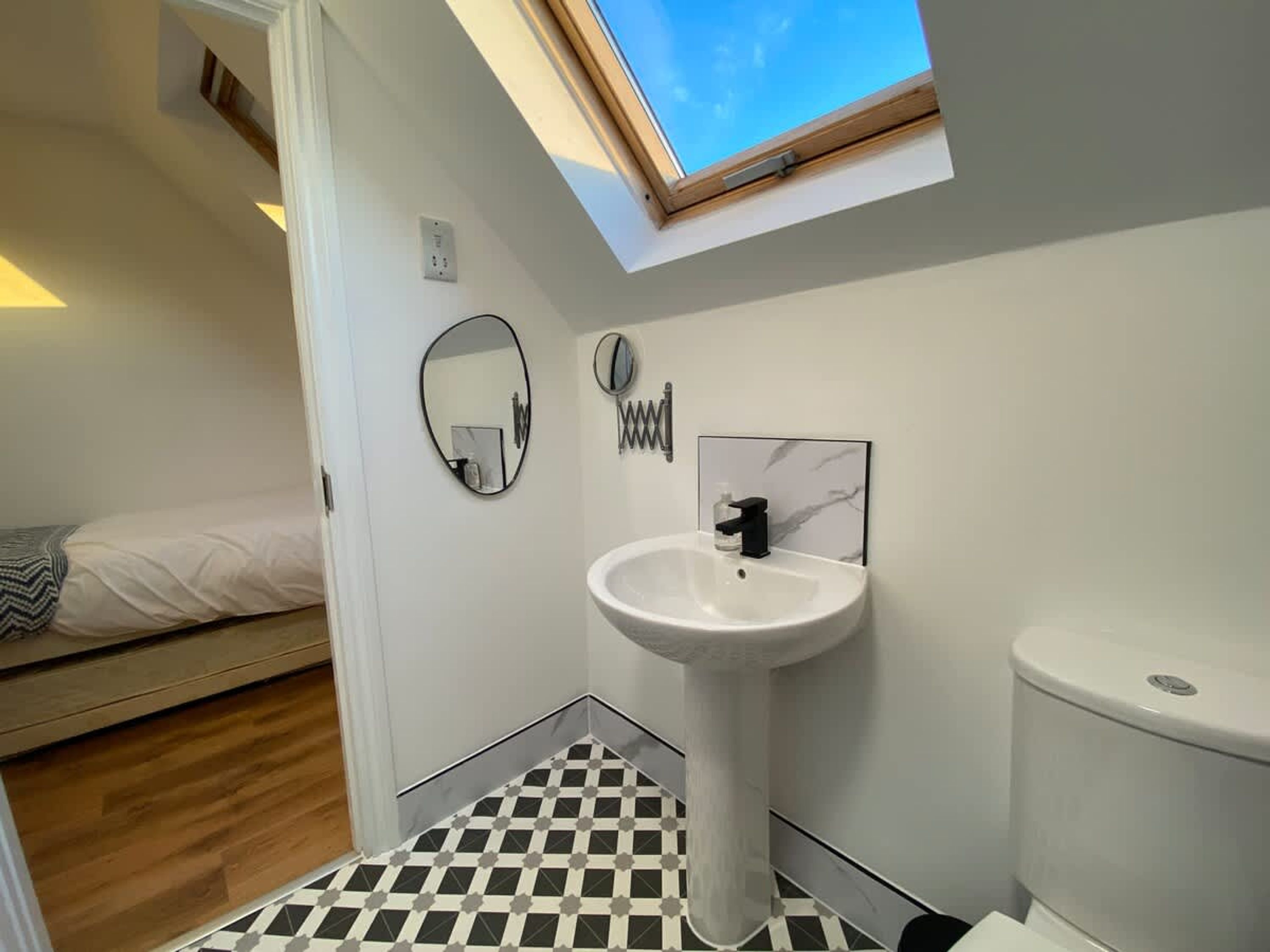 Une chambre à coucher de taille modeste, comprenant un lit, une lucarne, une salle de bain avec un lavabo et des toilettes, et un sol à carreaux.