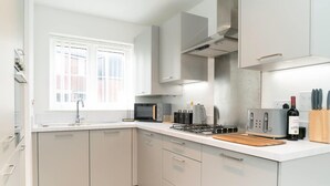 A modern kitchen with white cabinets, a gas stove, a microwave, and a window with blinds. - Host Stay Hewitt Close (Peterborough)