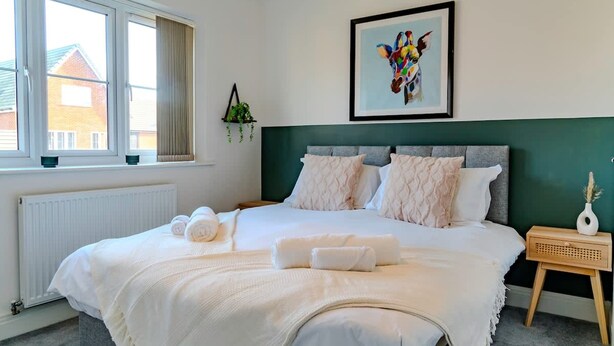 A neatly made bed with rolled towels, a framed artwork on the wall, and a window with a view of buildings outside. - Host Stay Hewitt Close (Peterborough)