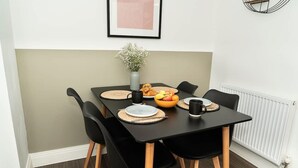 A dining area with a black table, four black chairs, a vase with flowers, a fruit bowl, and a framed picture on the wall. - Host Stay Hewitt Close (Peterborough)